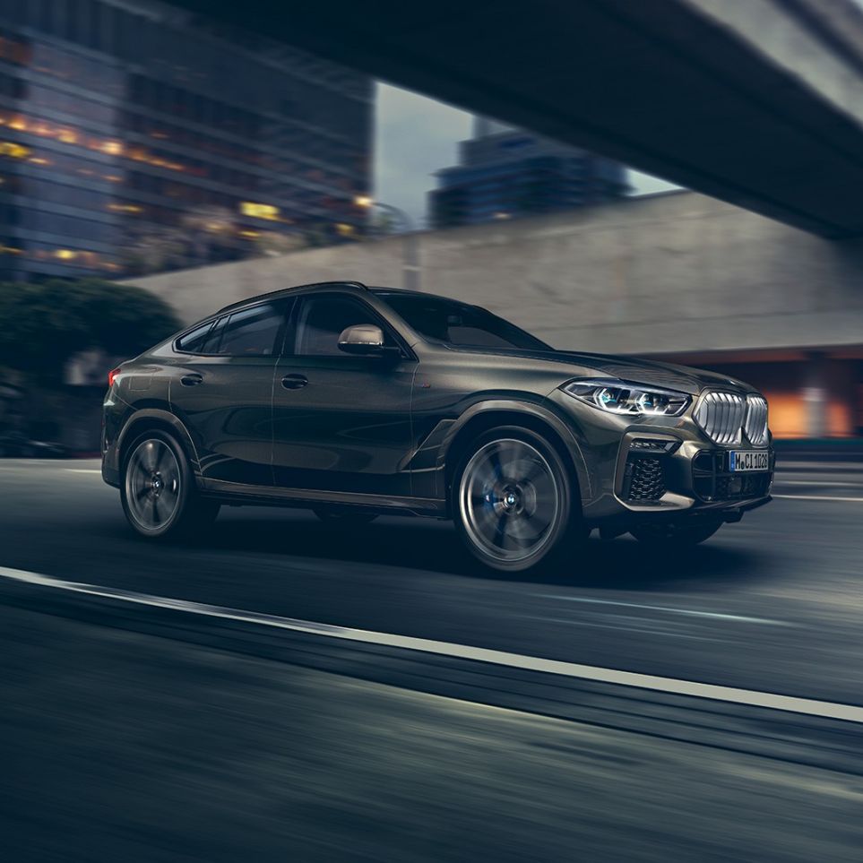 BMW X6 во движење. BMW X6 од тричетвртинска перспектива однапред во движење пред урбана заднина.
