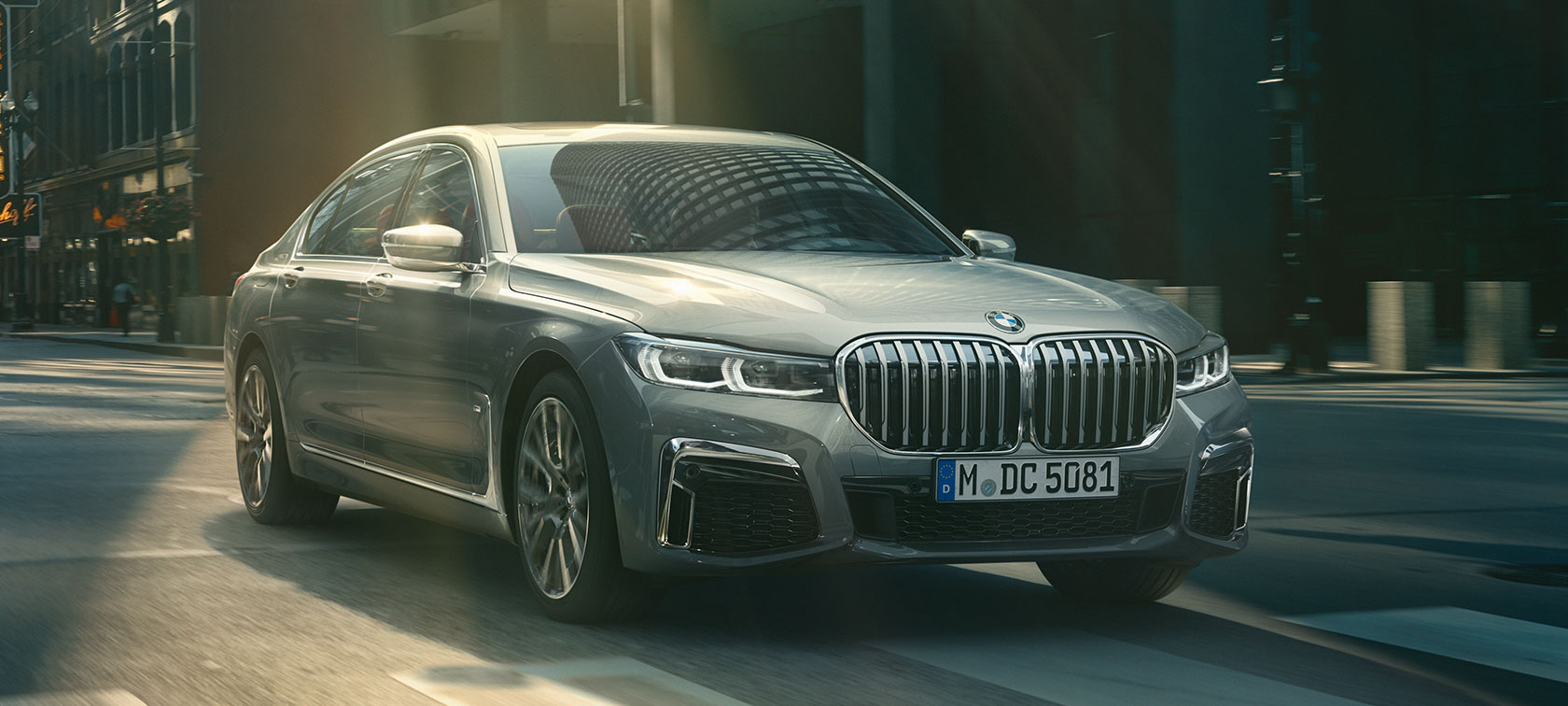 Сив BMW Серија 7 Седан во возење, тричетвртински преден кадар Сив BMW Серија 7 Седан вози низ урбана средина во тричетвртински преден кадар