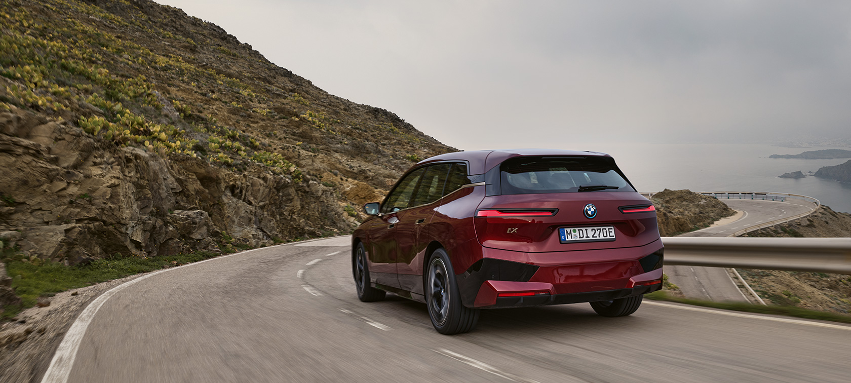 BMW iX 2021 електричен SUV BMW iX xDrive50вози по крајбрежен пат BMW iX 2021 електричен SUV BMW iX xDrive50 Aventurin Red црвена боја заден кадар од возење по крајбрежен пат