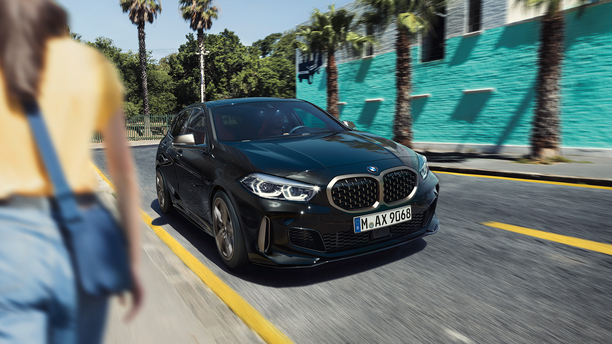 BMW M135i xDrive, тричетвртински приказ однапред. BMW M135i xDrive во Black Sapphire црна металик боја, тричетвртински приказ однапред со жена во предниот дел од сликата.