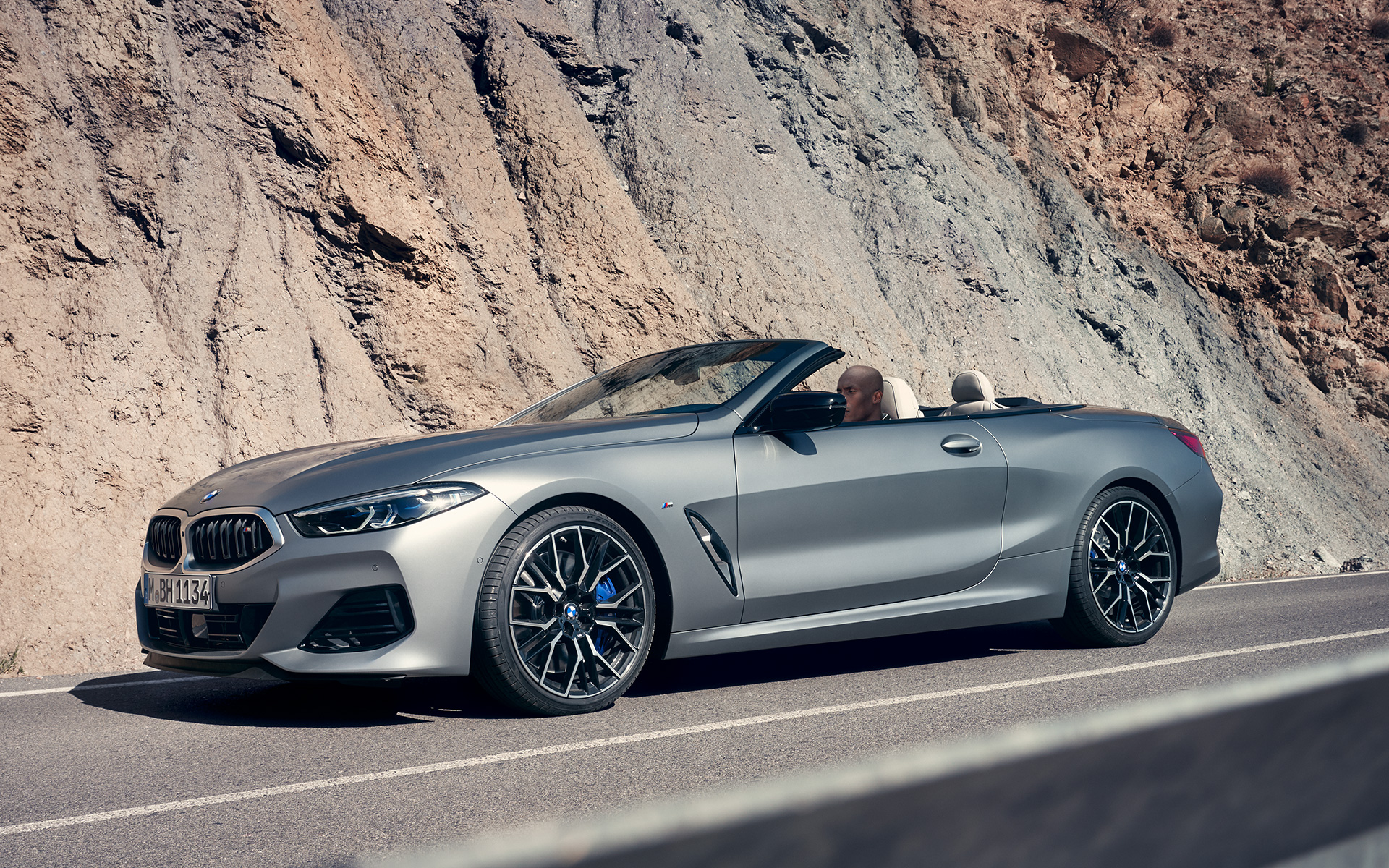 Тричетвртински поглед однапред на BMW M850i xDrive кабриолет G14 LCI Facelift 2022 BMW M850i xDrive кабриолет G14 LCI Facelift 2022 BMW Individual Frozen Pure Grey металик боја, тричетвртински поглед однапред во возење покрај карпест предел
