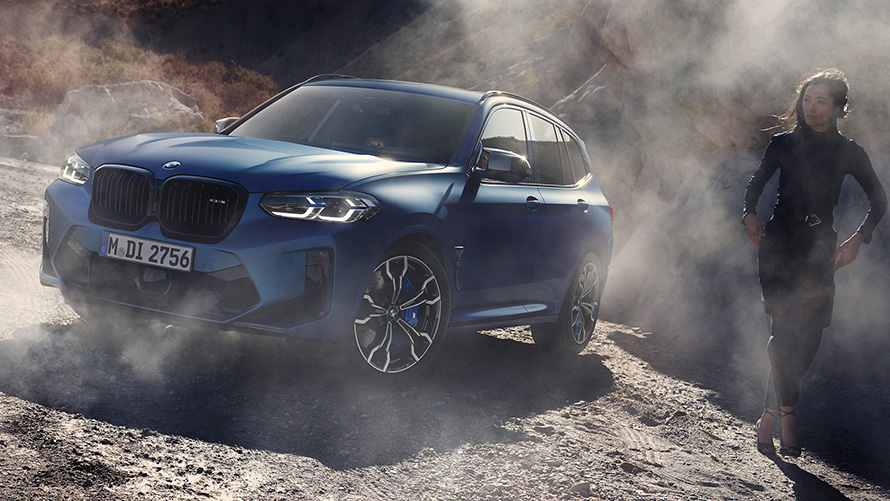 BMW X3 M Competition F97 LCI 2021 тричетвртински фронтален поглед во стоење на каменест терен BMW X3 M Competition F97 LCI Facelift 2021 Frozen Marina Bay сина металик боја тричетвртински фронтален поглед во стоење на каменест терен со жена модел.