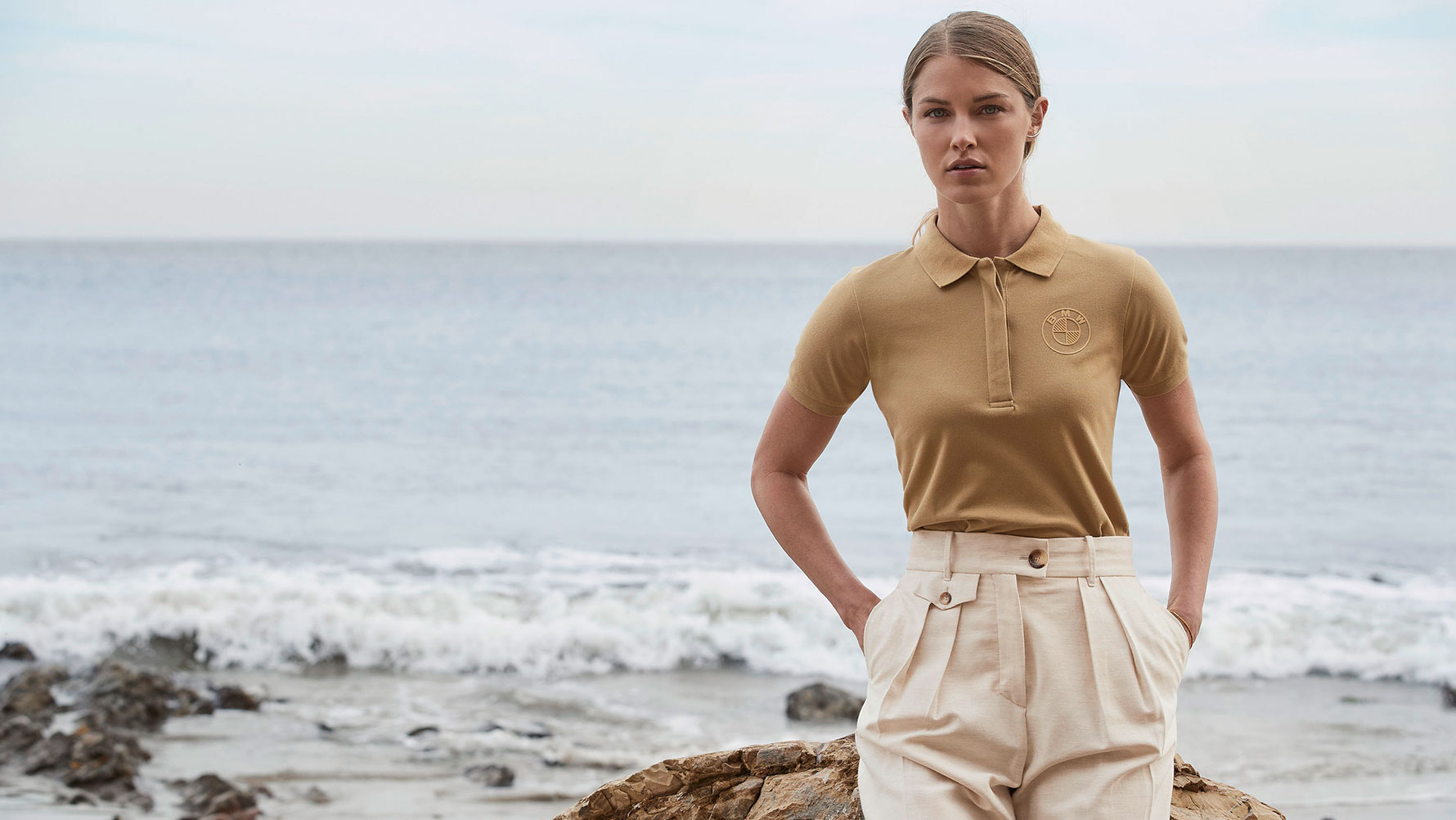 A young woman is wearing the BMW Polo Shirt Logo for ladies in front of a beach scenery. A young woman is wearing the BMW Polo Shirt Logo for ladies in front of a beach scenery.